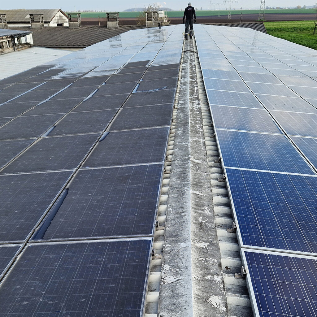 Solarreinigung in Aktion: Person steht auf Dach und bedient Solarreinigungsroboter. Auf einer Seite sind gereinigte Solarmodule, auf der anderen Seite müssen die PV-Module noch gereinigt werden. Die Module werden von SoluShine Solarreinigung gereinigt